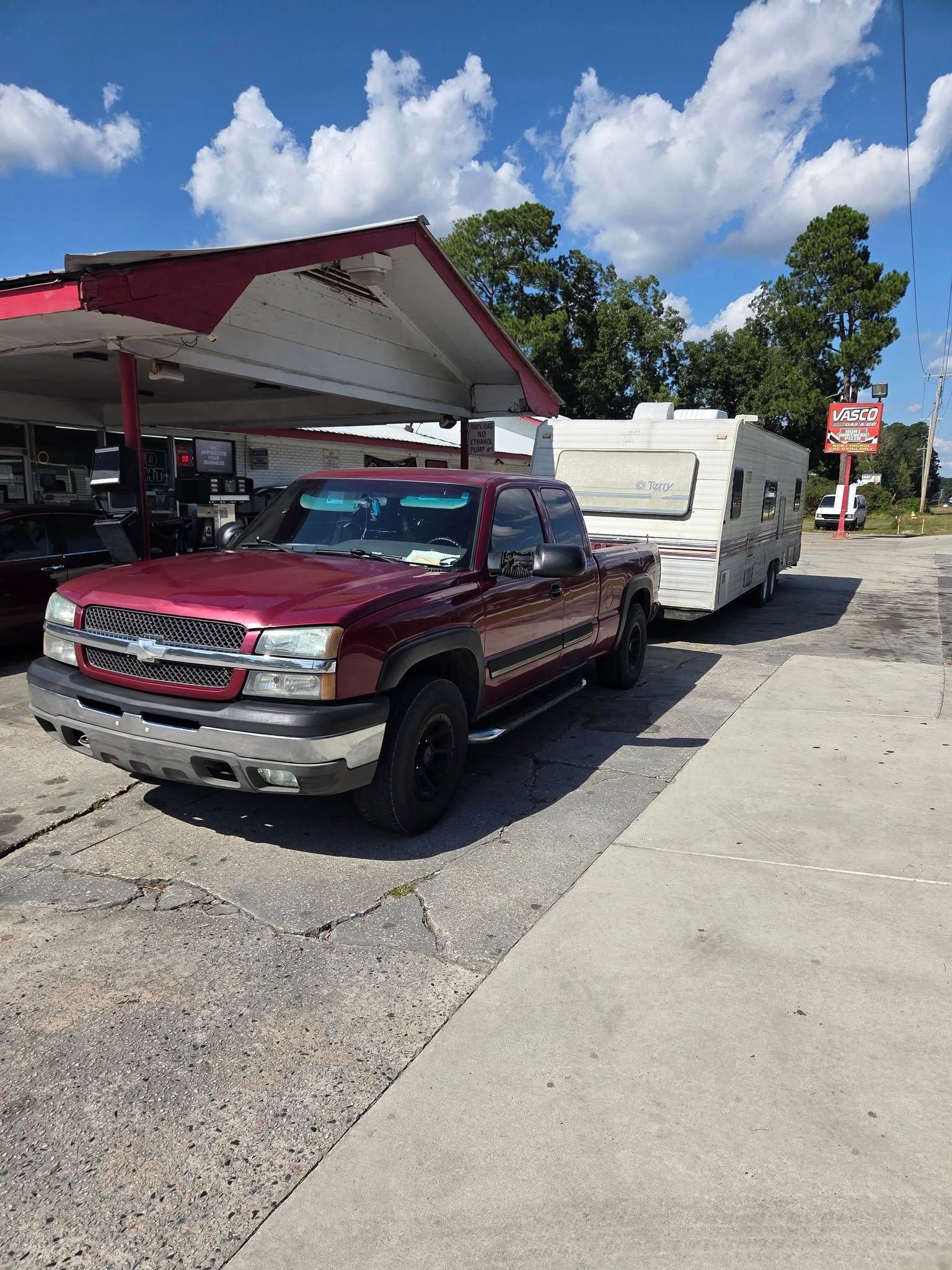 Maroon pickup truck towing a white camper parked beside a gas station under a blue sky