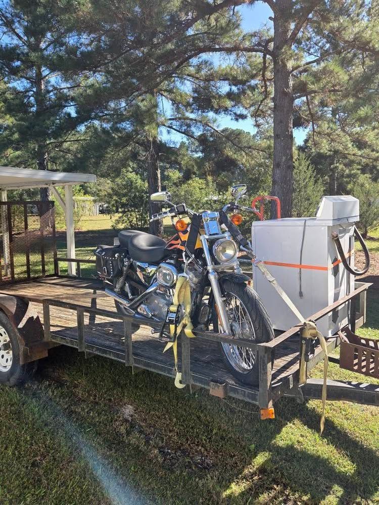 Motorcycle loaded on a flatbed trailer beside a house, with trees and a yard in the background