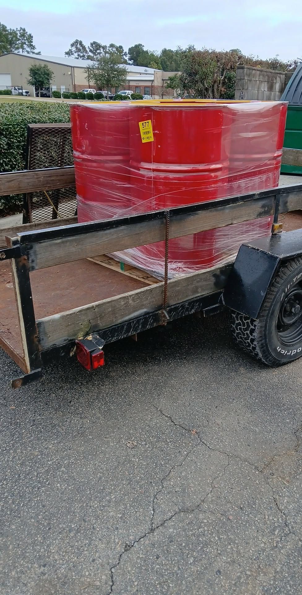 Red shrink-wrapped barrel secured on a black utility trailer in a parking lot.