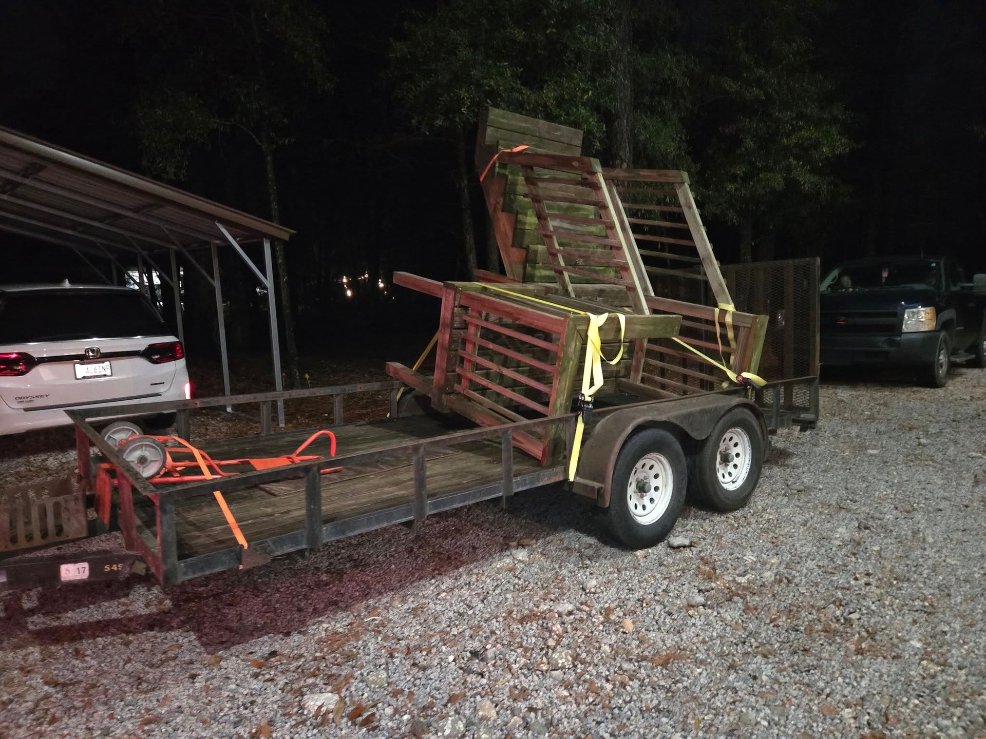 Red utility trailer with folded ramps and orange straps parked on gravel at night near a shed.