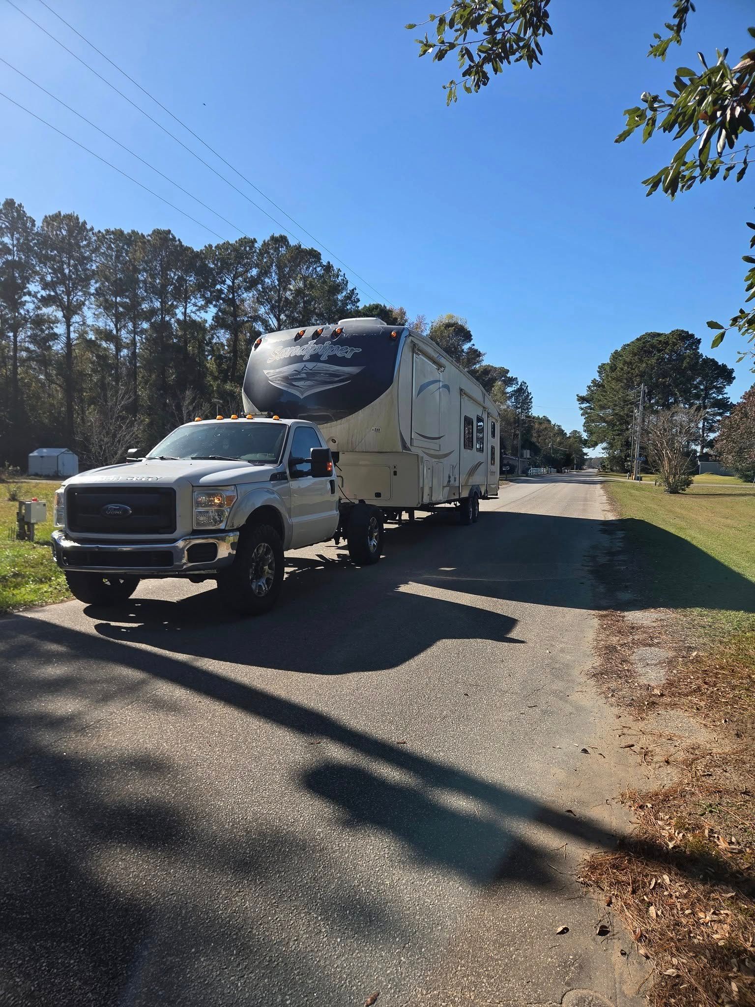 White RV and tow truck parked on a sunny rural road with trees and blue sky.