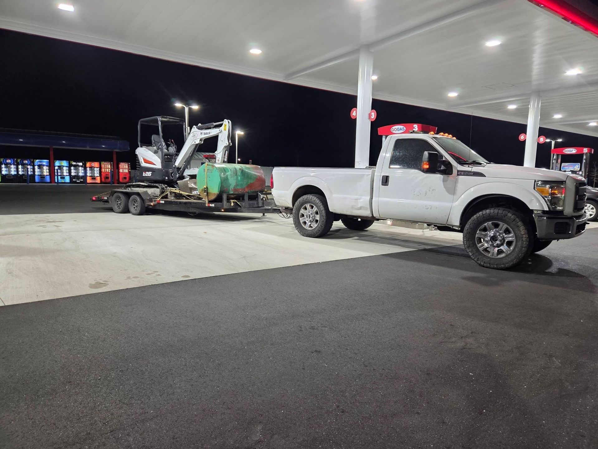 White pickup truck at a gas station loading a green dumpster onto a trailer at night