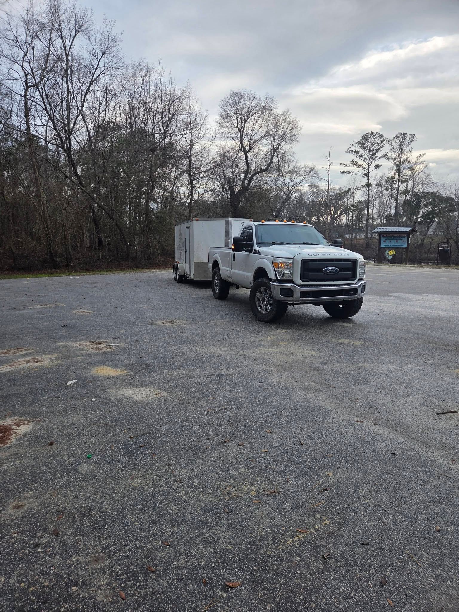 White pickup truck towing a white enclosed trailer in a gravel lot beside leafless trees
