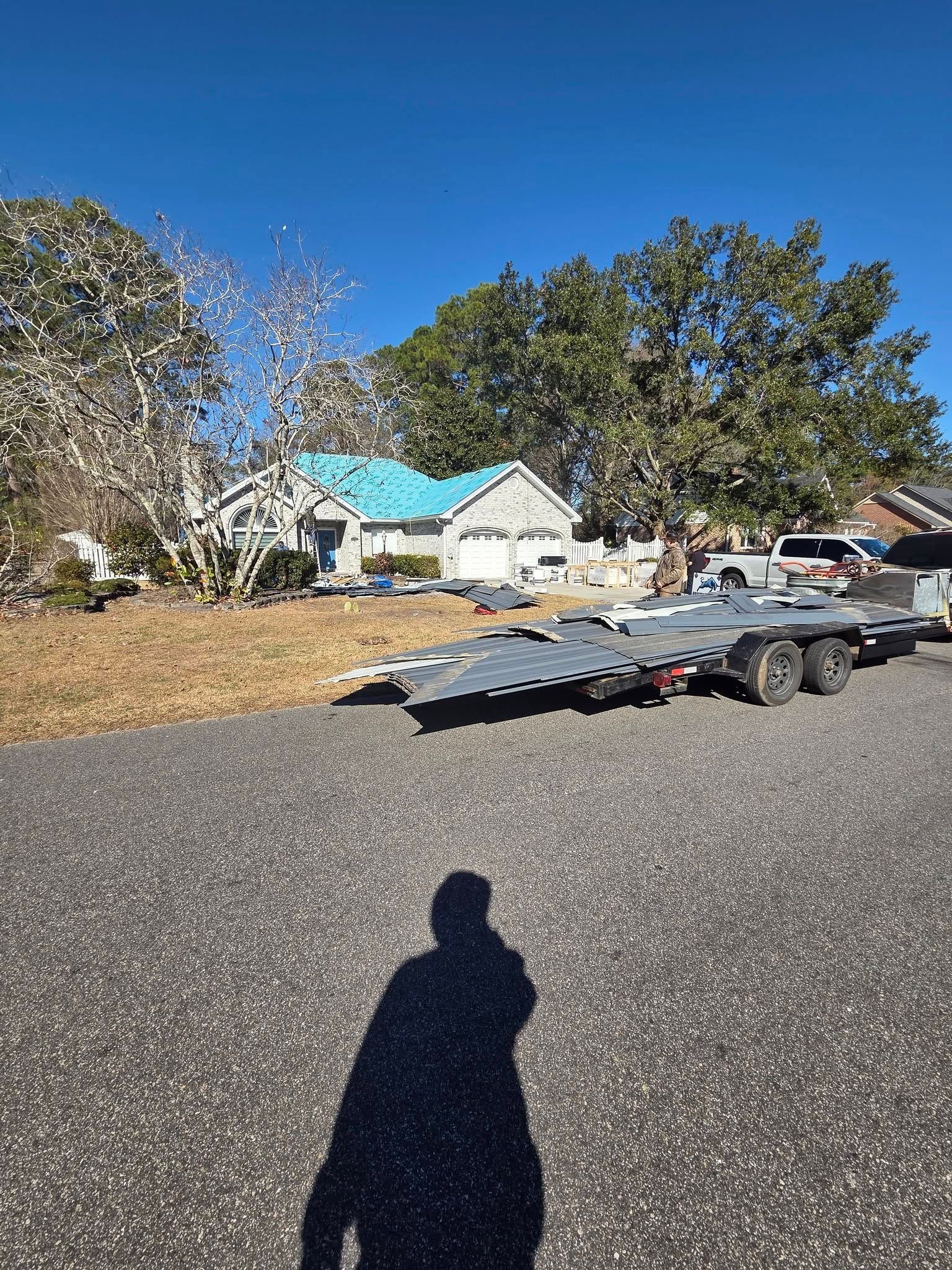 House with turquoise roof behind trees, parked trailers in a gravel driveway, and a clear blue sky.