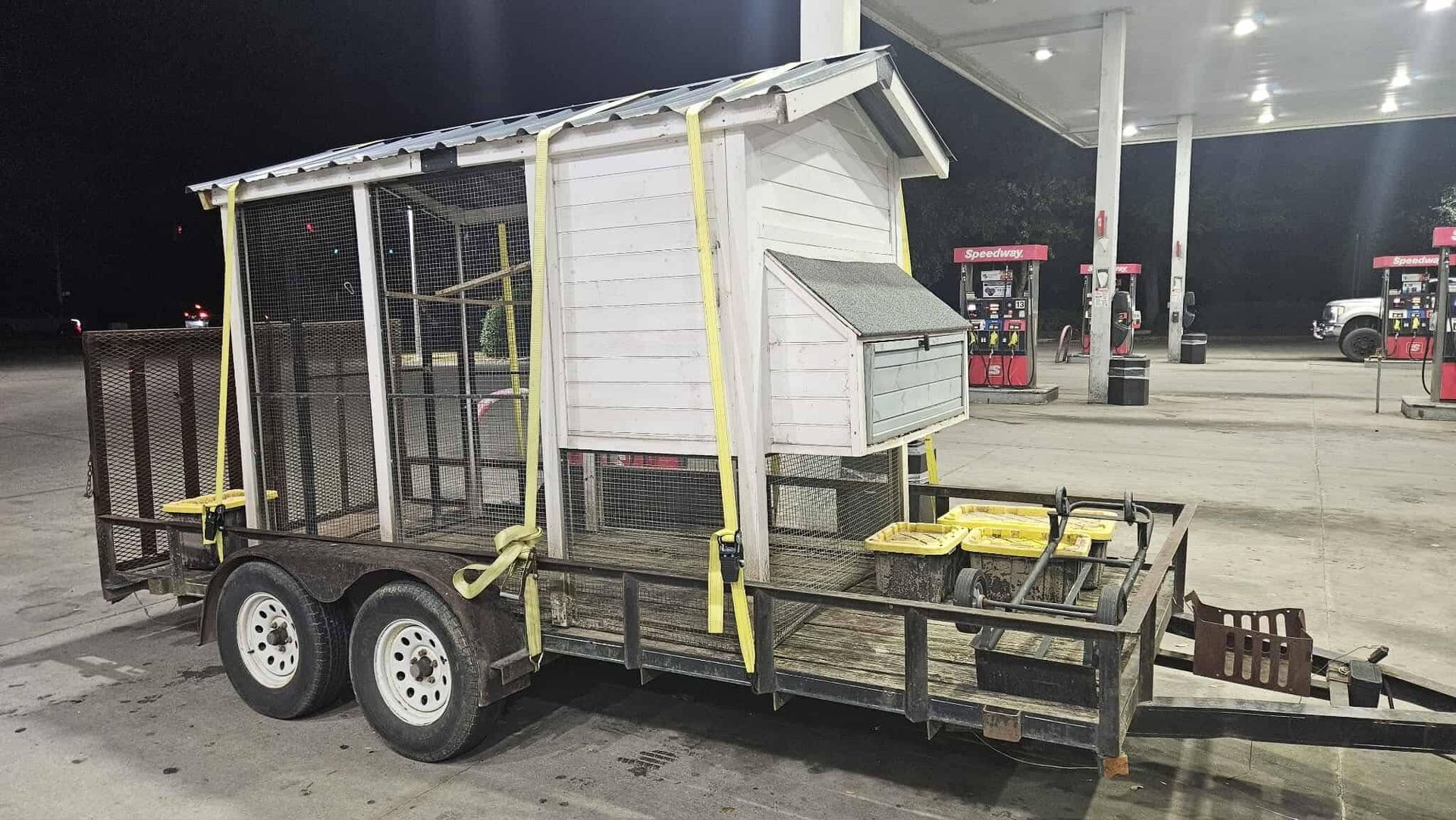 Small white utility trailer with metal cage, yellow straps, and a front hitch parked at a gas station at night