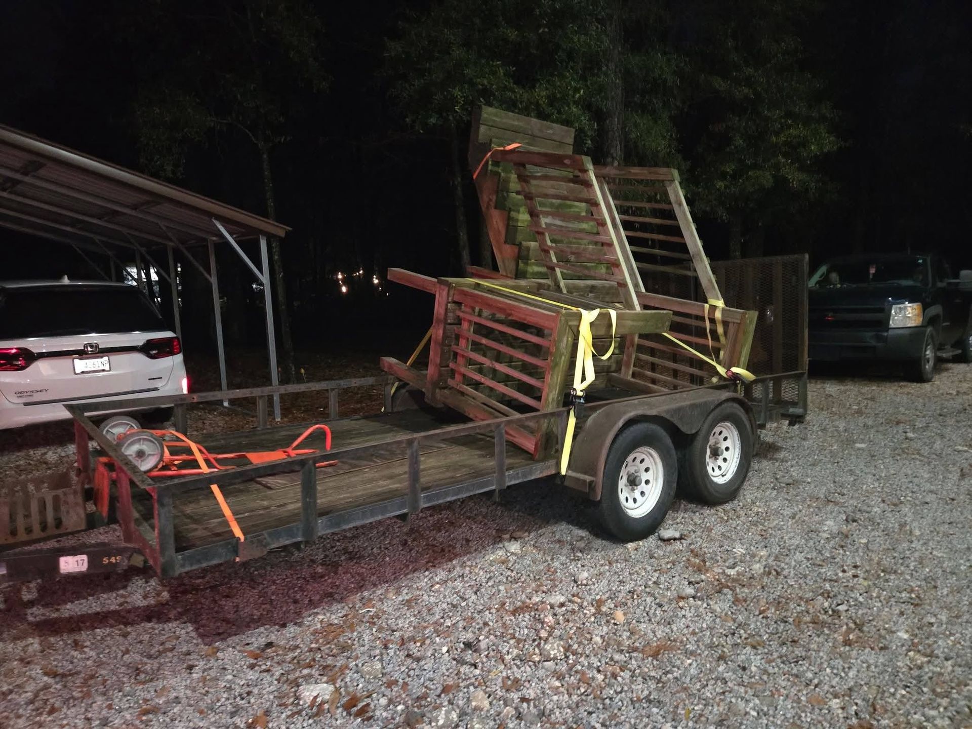 Tilt trailer loaded with wooden pallets and metal racks on a gravel lot at night