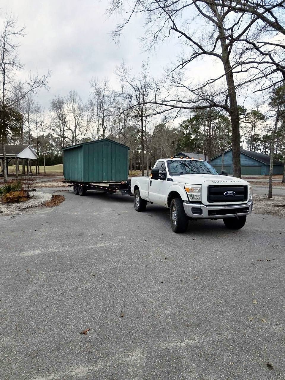 White pickup truck towing a green shed on a trailer in a parking lot with bare trees.