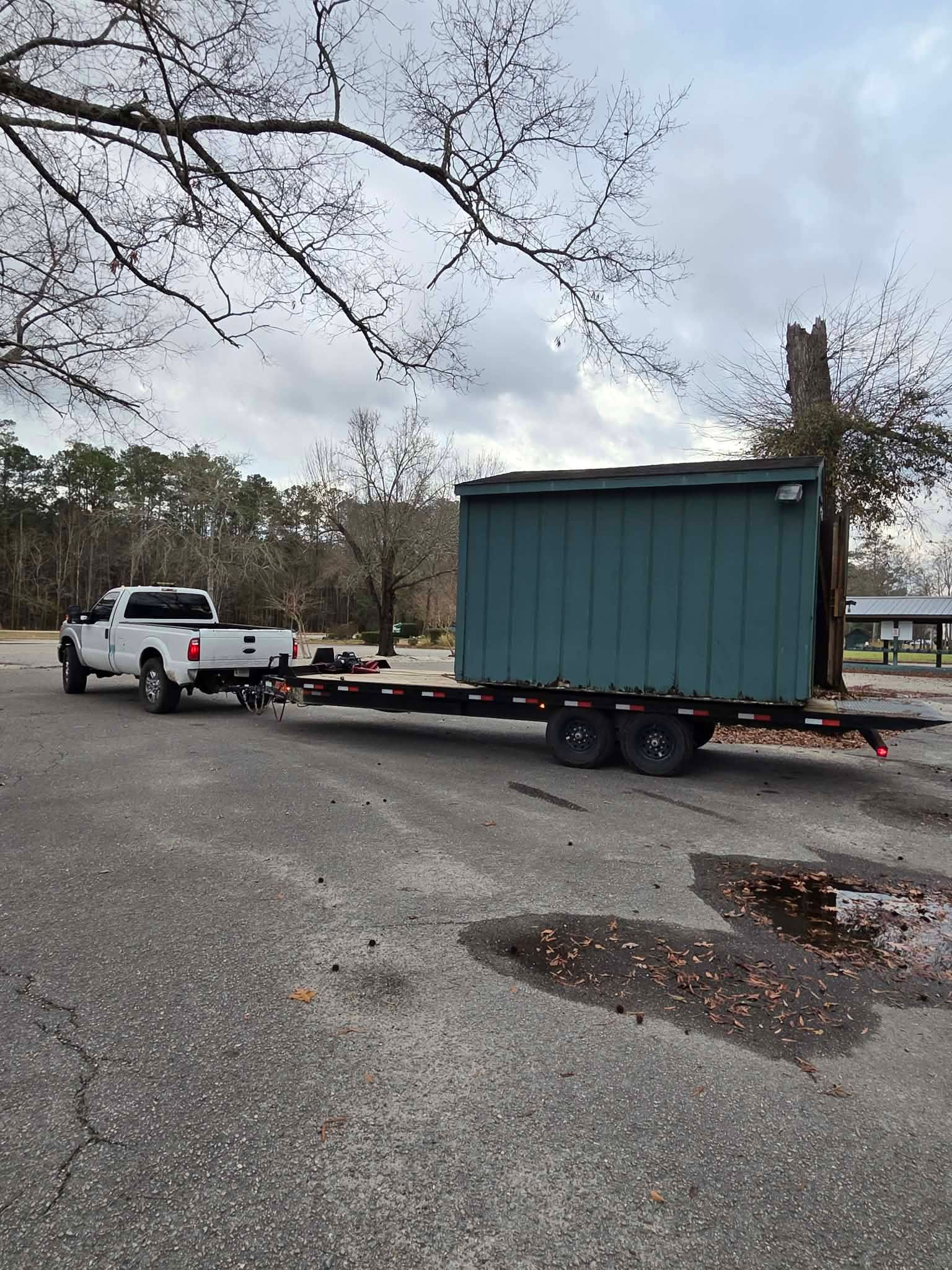 White pickup towing a green shed on a trailer in a gravel parking area under bare trees