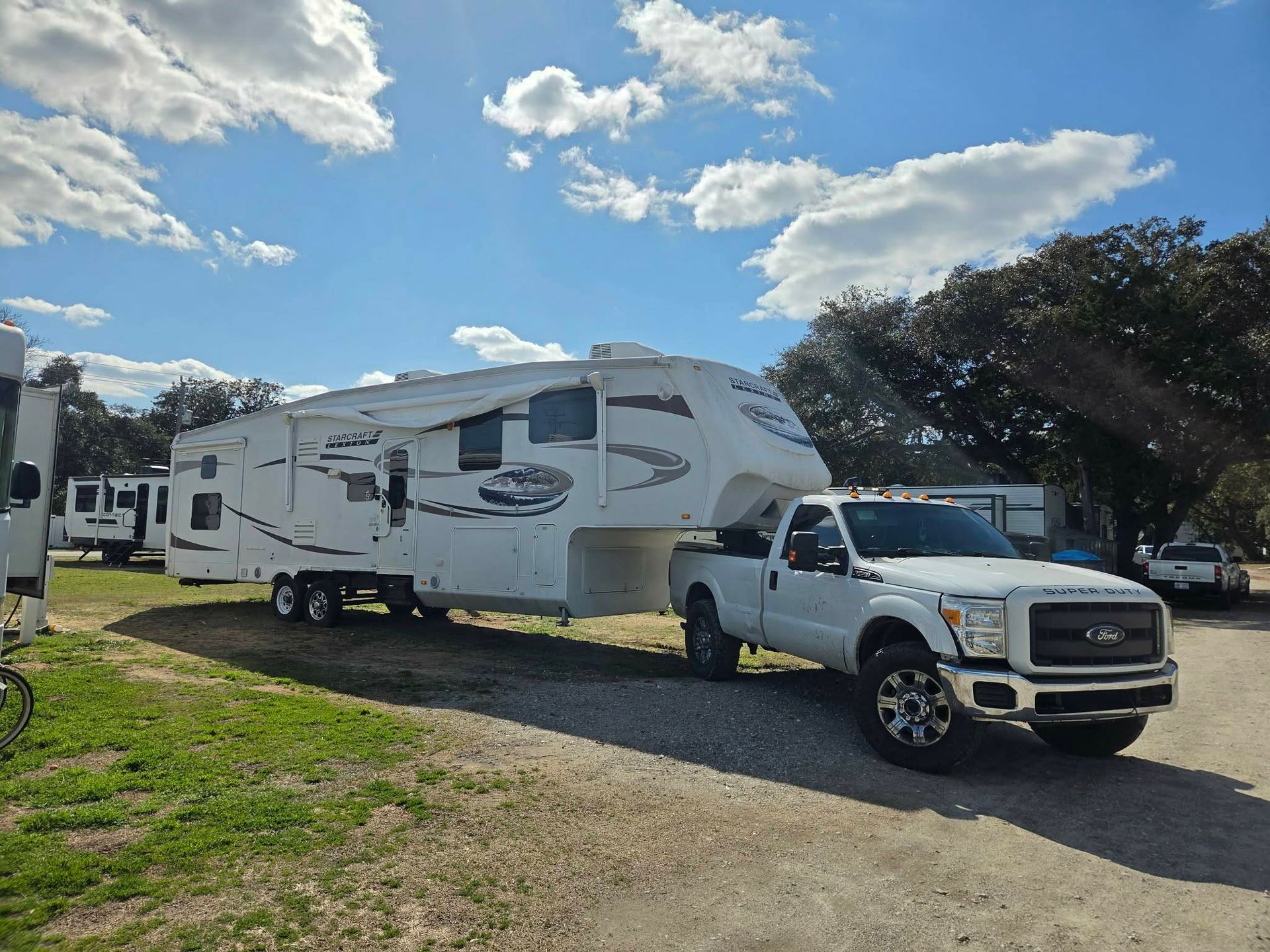 White pickup truck towing a large white camper trailer on a grassy lot under a partly cloudy sky