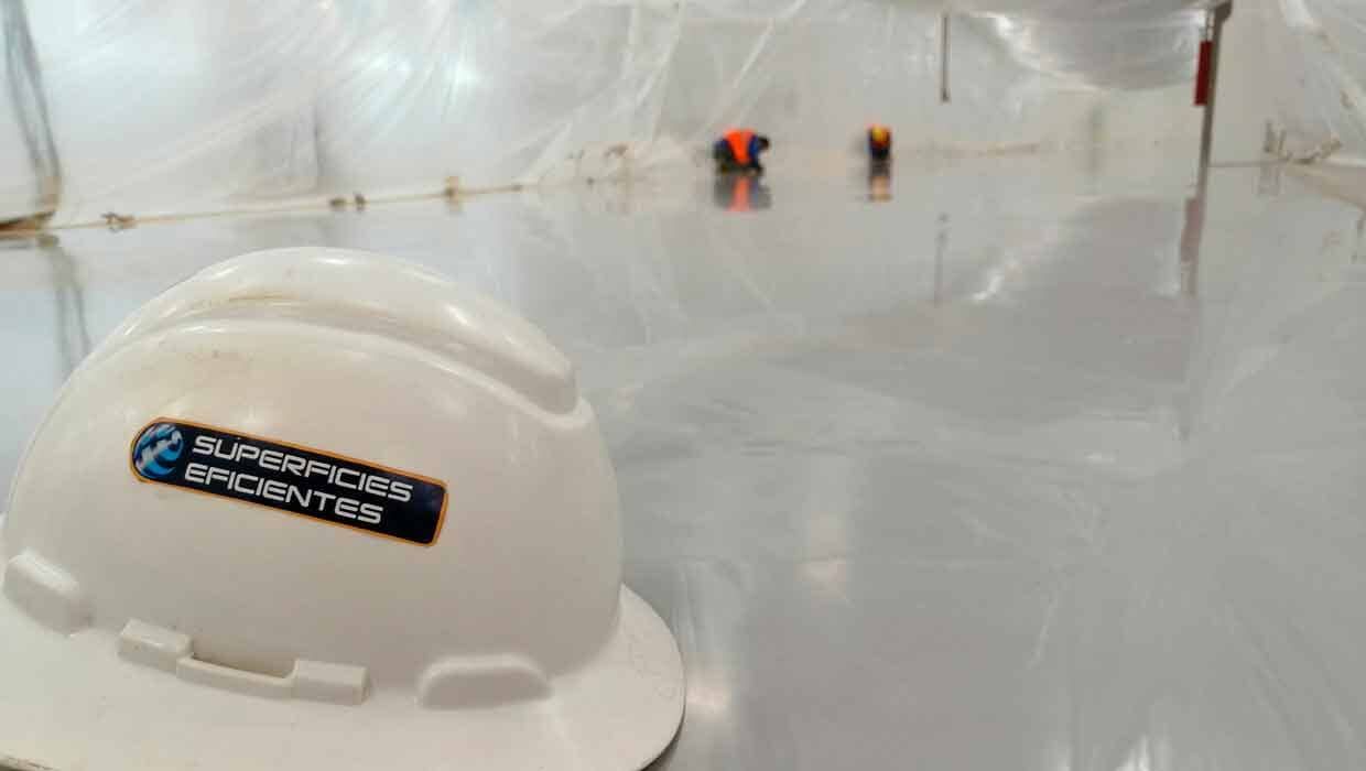 Un casco blanco se ve en primer plano mientras los trabajadores aplican un revestimiento de suelo liso, brillante y reflectante de color claro.