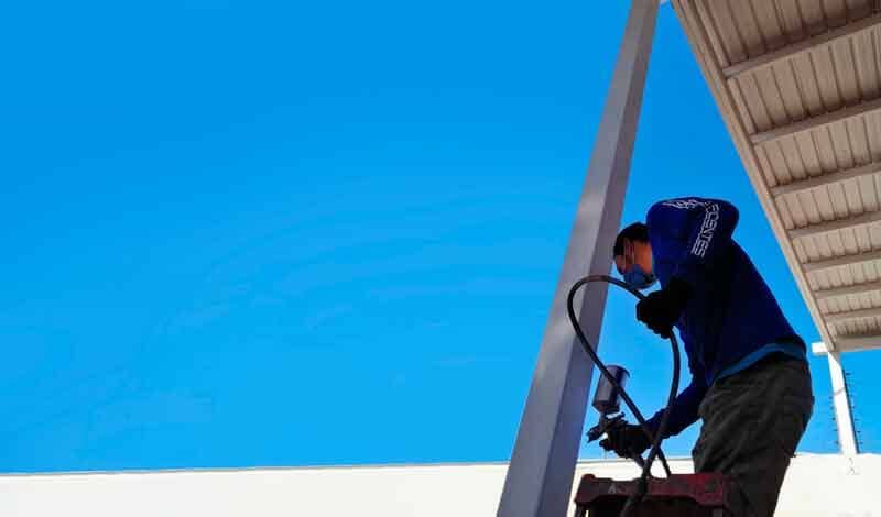 Un trabajador con una camisa azul de manga larga y pantalones oscuros utiliza una pistola pulverizadora de pintura sobre un poste de metal bajo un cielo azul despejado.