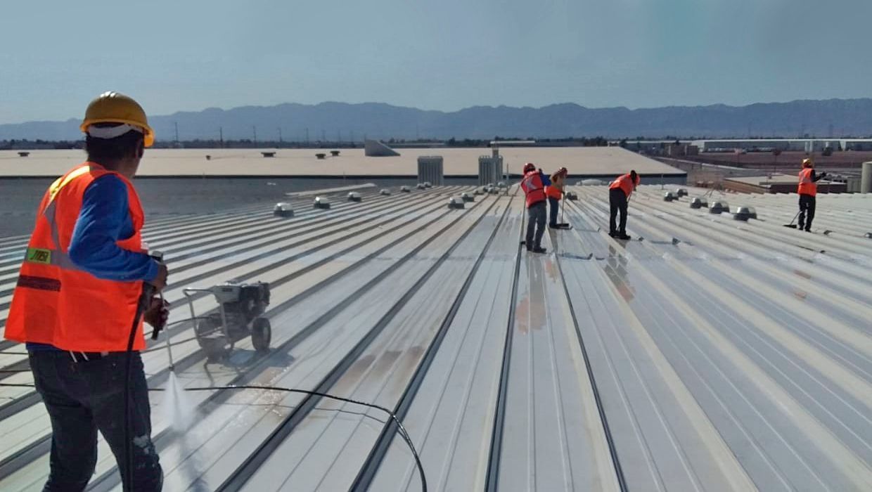 Trabajadores con chalecos de seguridad naranjas y cascos limpian con agua a presión y preparan un gran techo metálico industrial bajo un cielo despejado.