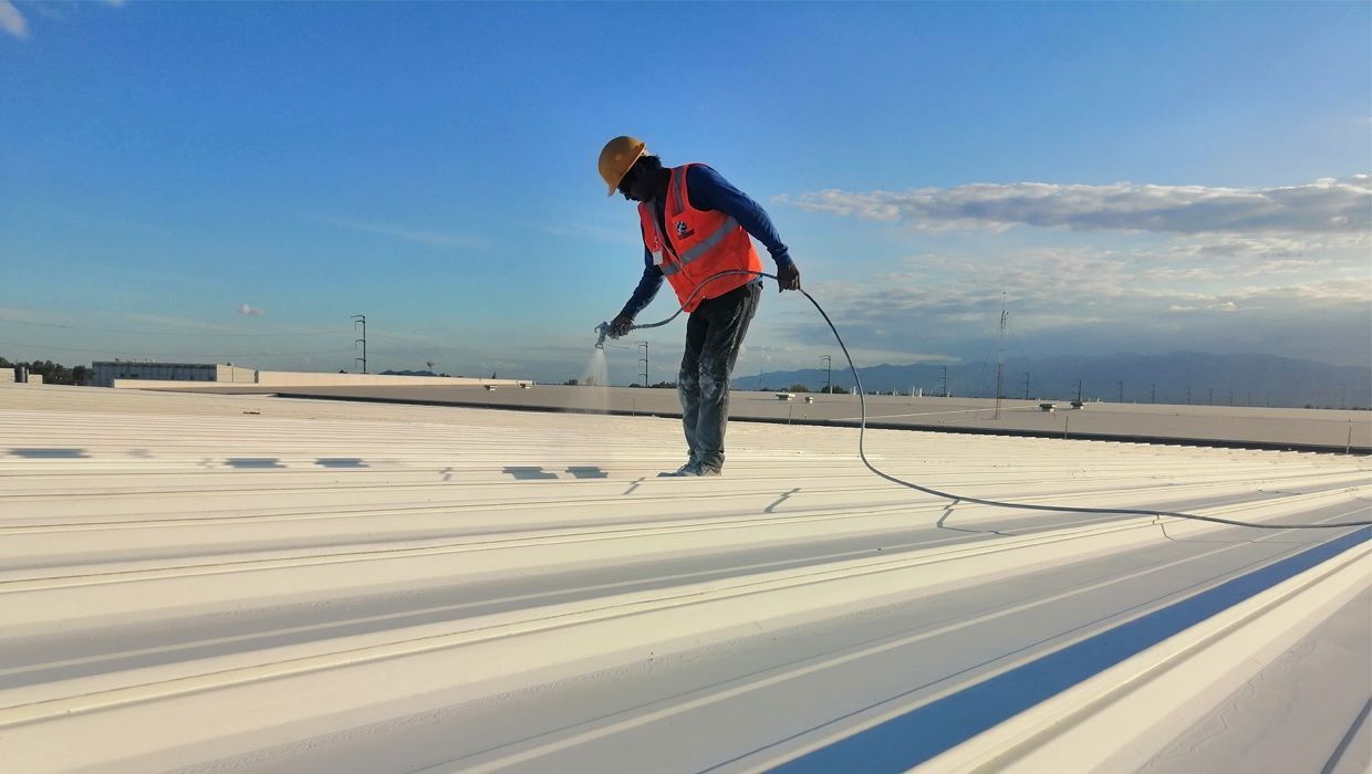 Un trabajador con chaleco de seguridad utiliza una herramienta pulverizadora para aplicar sellador blanco a un techo industrial plano bajo un cielo azul brillante.