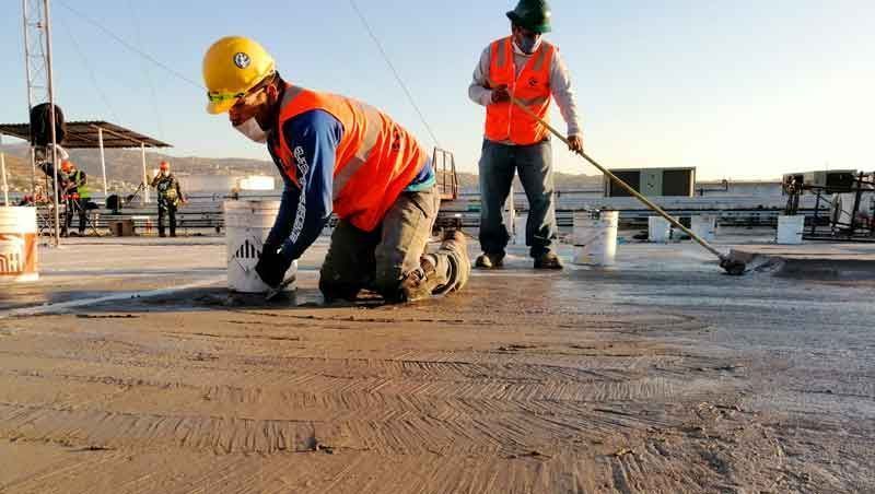 Dos obreros de la construcción, con chalecos reflectantes y cascos, aplican un sellador a un tejado bajo un cielo despejado.