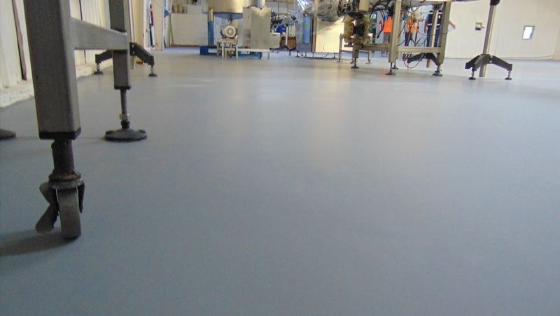 Un suelo industrial liso de color gris claro en el interior de unas instalaciones, con patas de equipos metálicos y trabajadores al fondo.