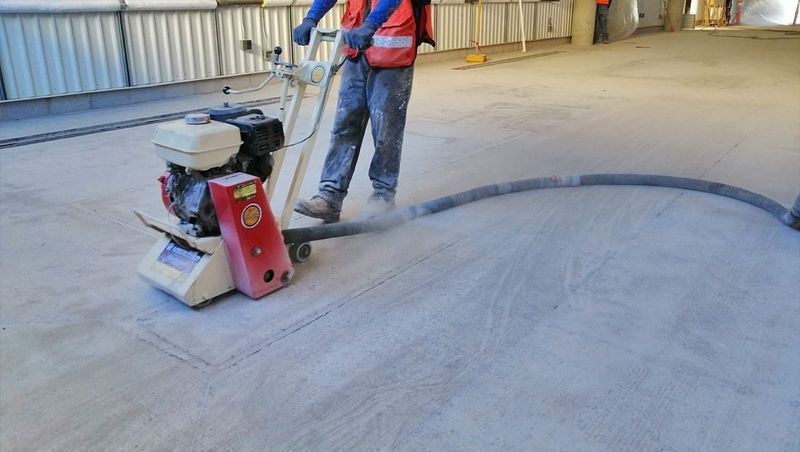 Un trabajador con un chaleco rojo maneja una pulidora de pisos de concreto en una obra de construcción, con una manguera conectada para la extracción de polvo.