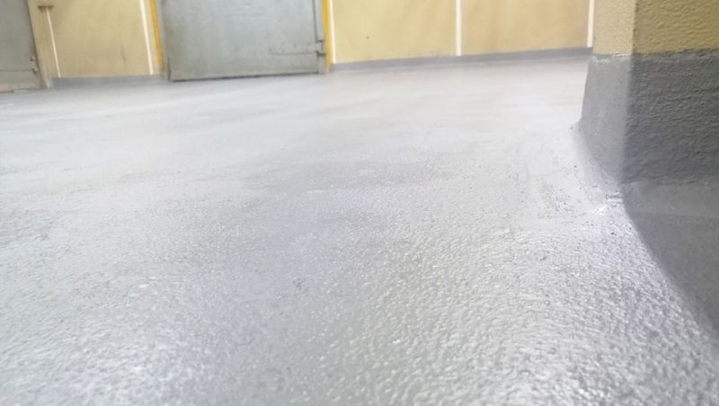 Primer plano de un revestimiento de suelo de epoxi gris recién aplicado en una habitación con una pared en esquina y el marco de una puerta al fondo.