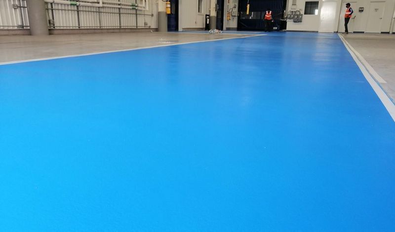 Una amplia zona interior con un suelo liso de resina epoxi de color azul brillante, delimitada por líneas blancas pintadas, con gente a lo lejos.