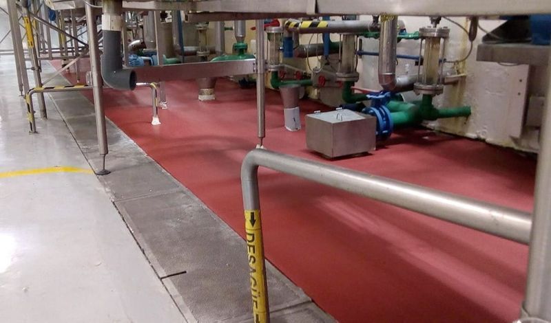 Suelo de una instalación industrial con una sección pintada de rojo, patas de maquinaria metálicas, tuberías y marcas de seguridad amarillas.