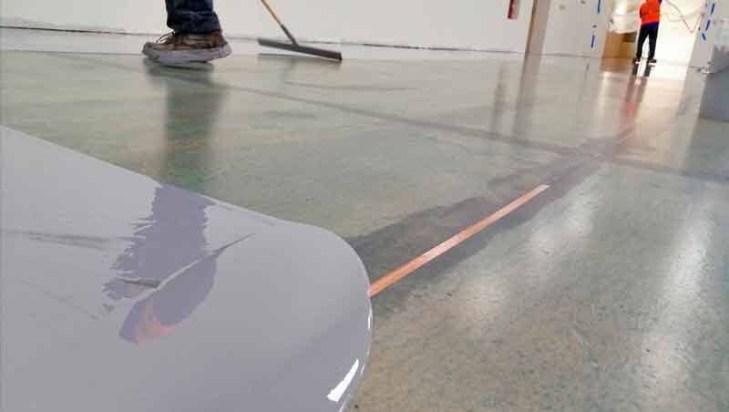 En un espacio comercial, los trabajadores utilizan una espátula para extender una capa de revestimiento epoxi gris claro sobre un suelo de hormigón.