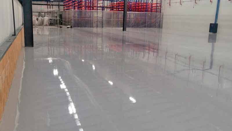 Un suelo liso de resina epoxi gris claro en un gran almacén moderno, con columnas de acero estructural y estanterías al fondo.