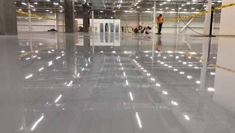 Trabajadores aplicando un acabado de epoxi gris brillante al suelo reflectante de un gran almacén.
