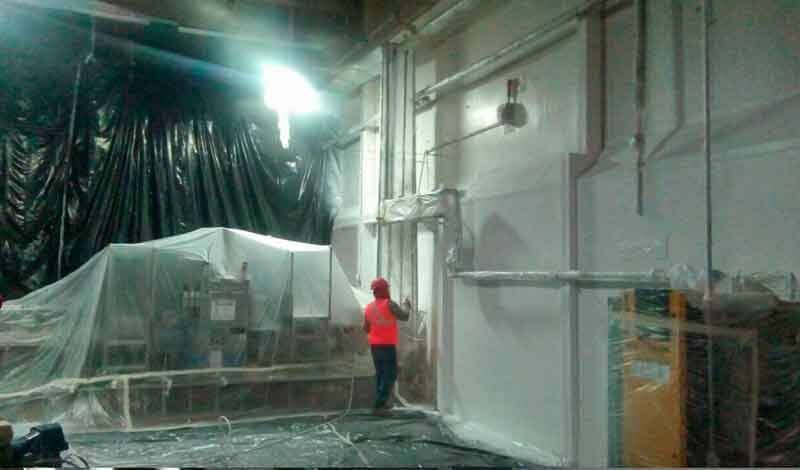 Una persona con un chaleco naranja de alta visibilidad rocía con pintura una pared industrial blanca dentro de una instalación cubierta con láminas de plástico.