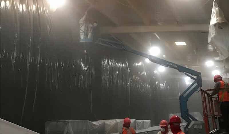 Trabajadores con cascos y chalecos protectores manejan una plataforma elevadora para alcanzar el techo dentro de una instalación industrial con poca luz.
