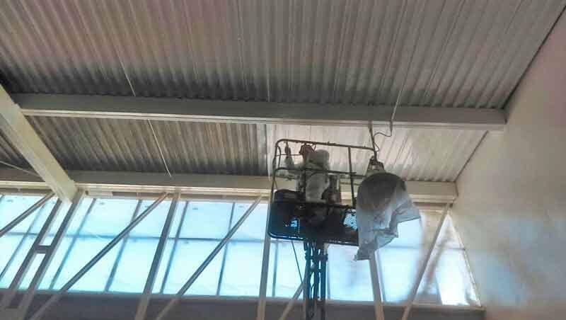 Un trabajador, desde la cesta de un ascensor, está pintando con aerosol el techo de chapa ondulada de un edificio industrial.