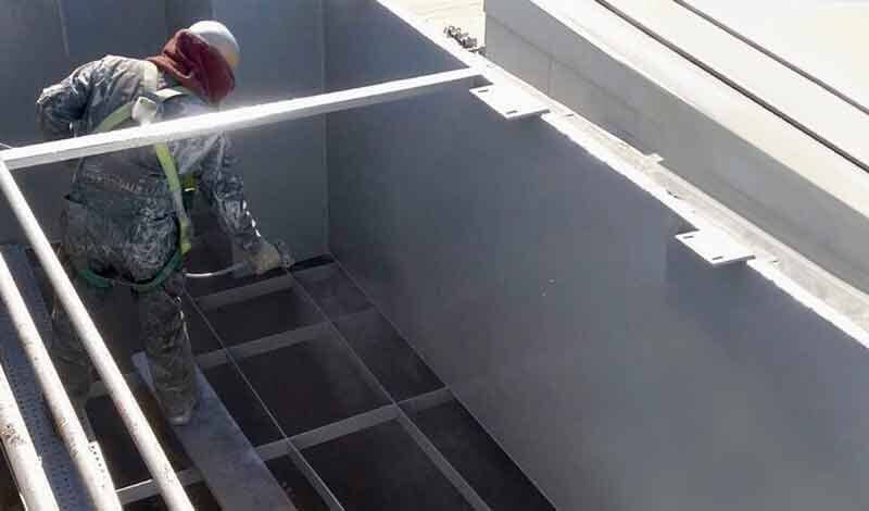 Un trabajador, con arnés y casco de seguridad, utiliza una pistola pulverizadora para aplicar una capa protectora de color gris a una estructura metálica.