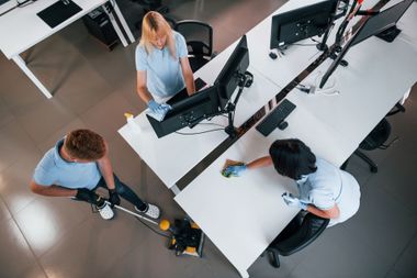 Three coworkers collaborate at a modern office desk with computer monitors and cables.