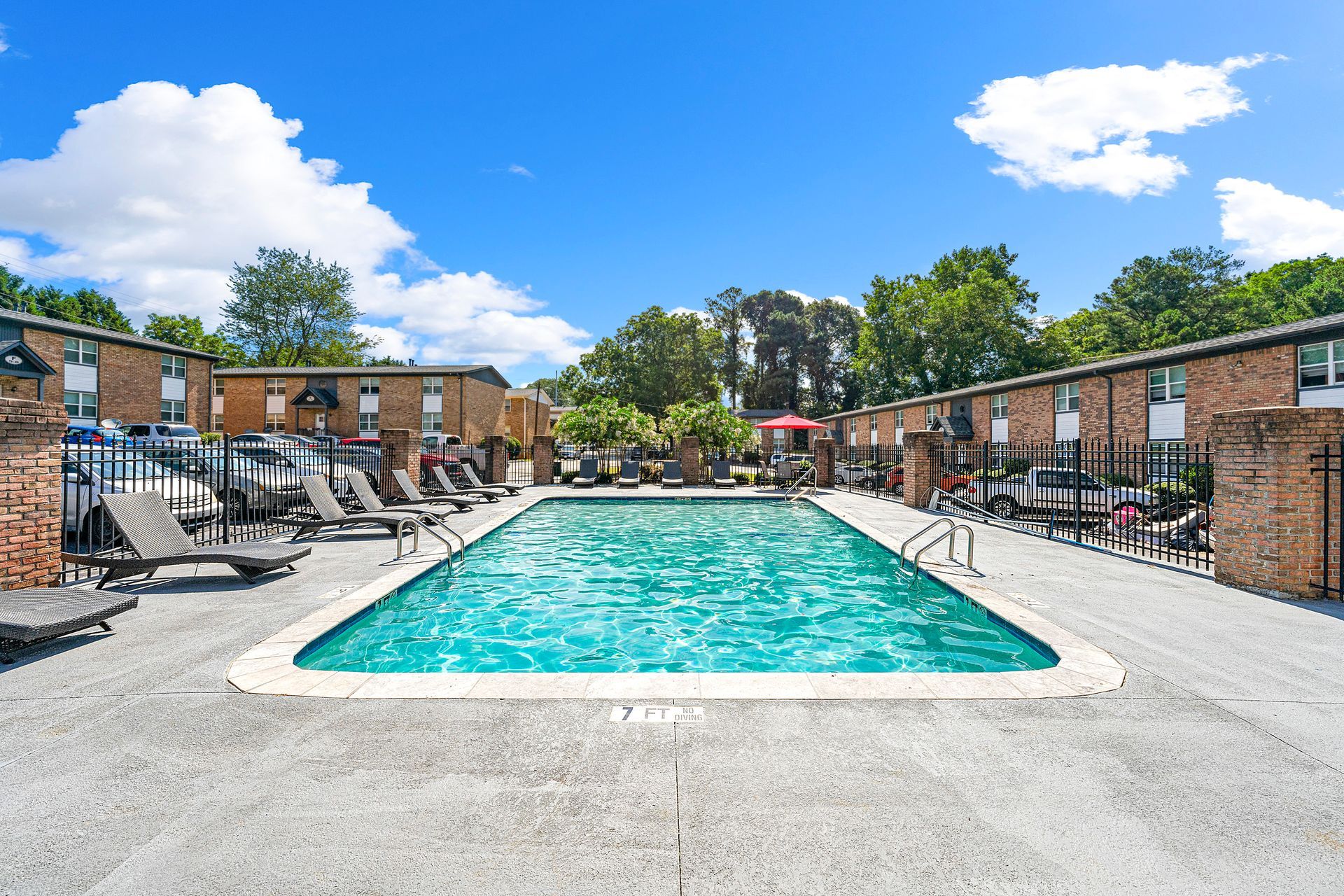 Outdoor apartment pool with turquoise water, lounge chairs, and brick buildings under a sunny blue sky