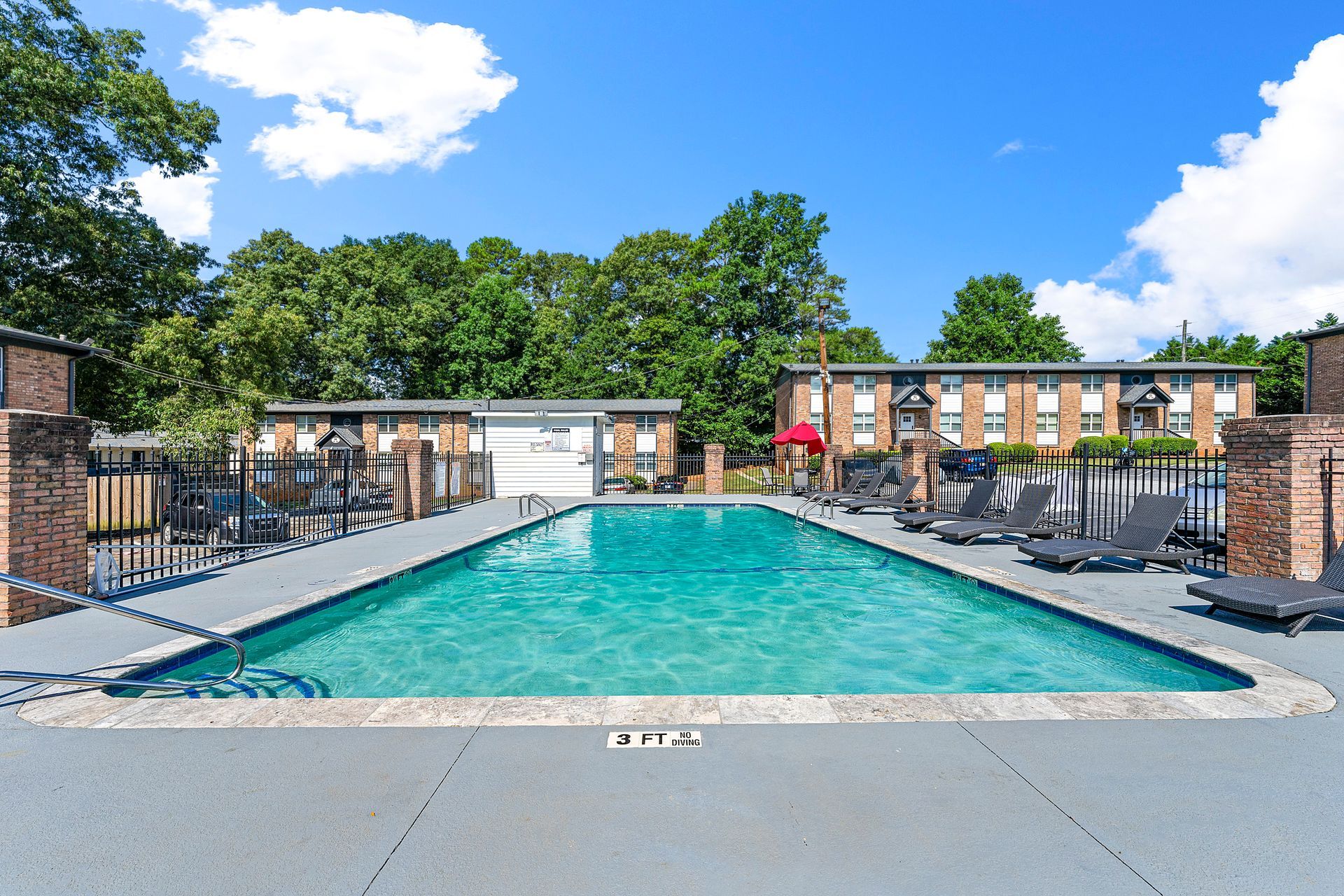 Outdoor apartment swimming pool with turquoise water, lounge chairs, and brick buildings under a blue sky