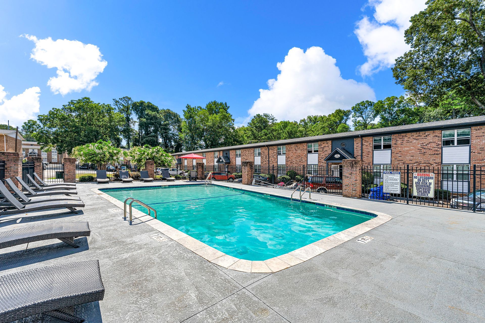 Apartment courtyard with turquoise swimming pool, lounge chairs, and brick buildings under a bright blue sky