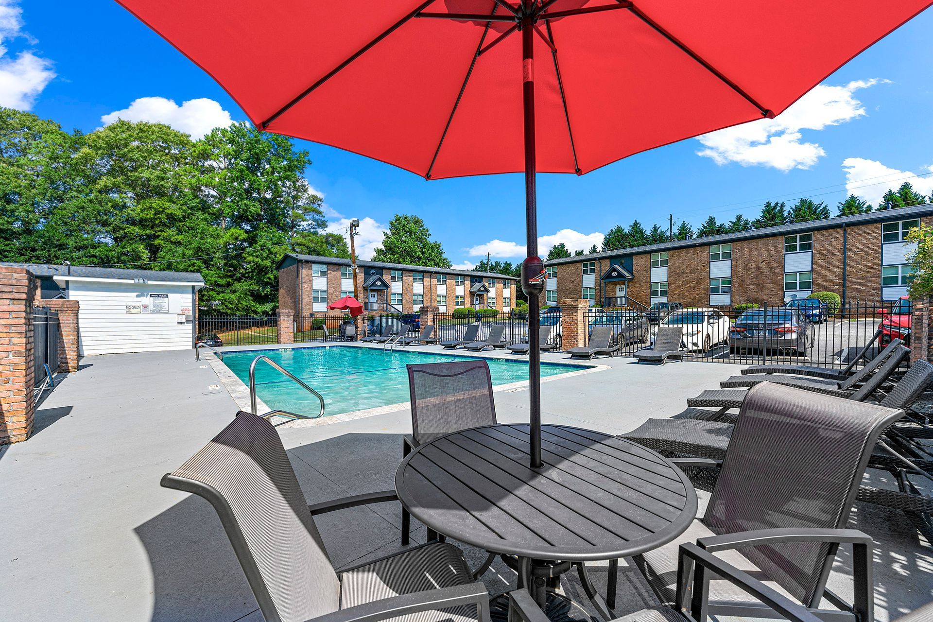 Red umbrella over a poolside patio with table and chairs, overlooking an outdoor swimming pool and apartment buildings