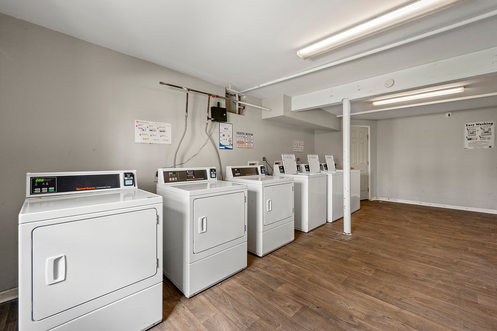 Laundry room with several white washing machines and dryers on a wood floor, under fluorescent lights