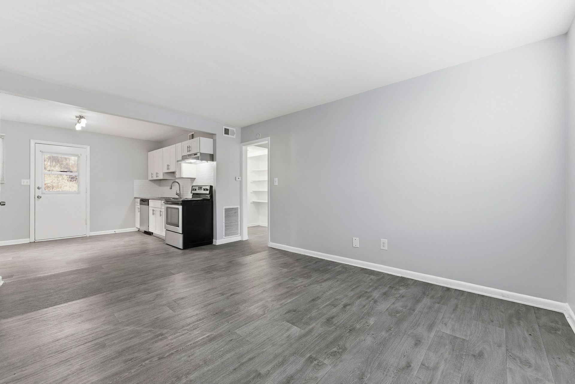 Empty modern apartment living area with gray floors, white walls, and a small kitchen.