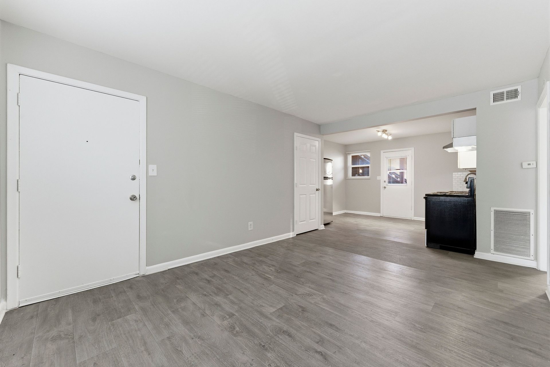 Empty gray living room with hardwood floors, white doors, and a view into a kitchen area