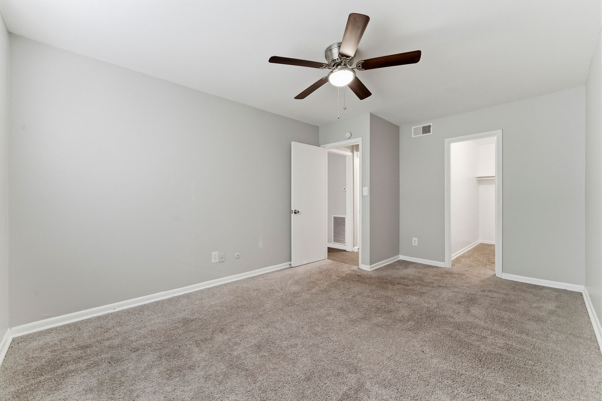 Empty carpeted bedroom with light gray walls, ceiling fan, and adjoining doorway to a hall and bathroom.