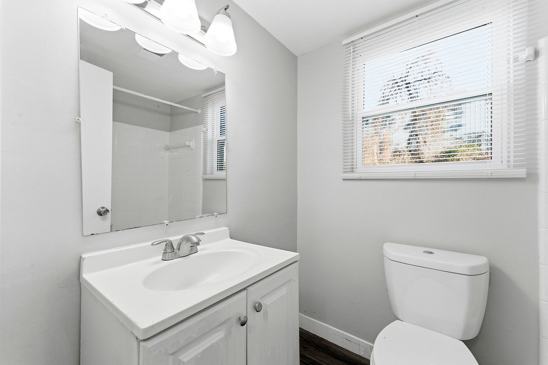 Small white bathroom with vanity, mirror, toilet, and a window with blinds.