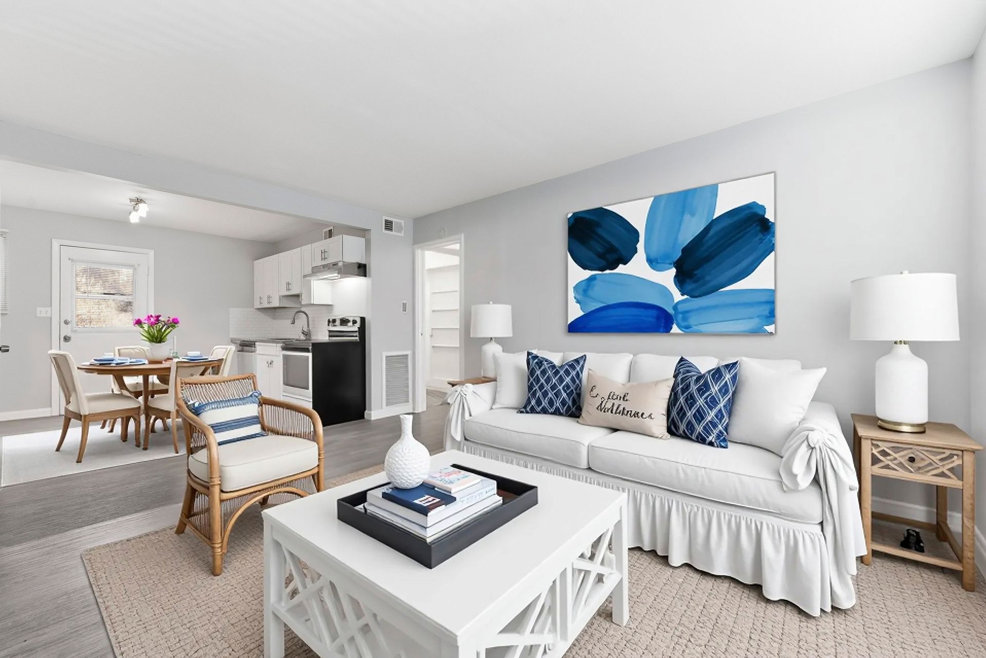 Bright living room with white sofa, blue abstract art, wicker chair, coffee table, and open dining area