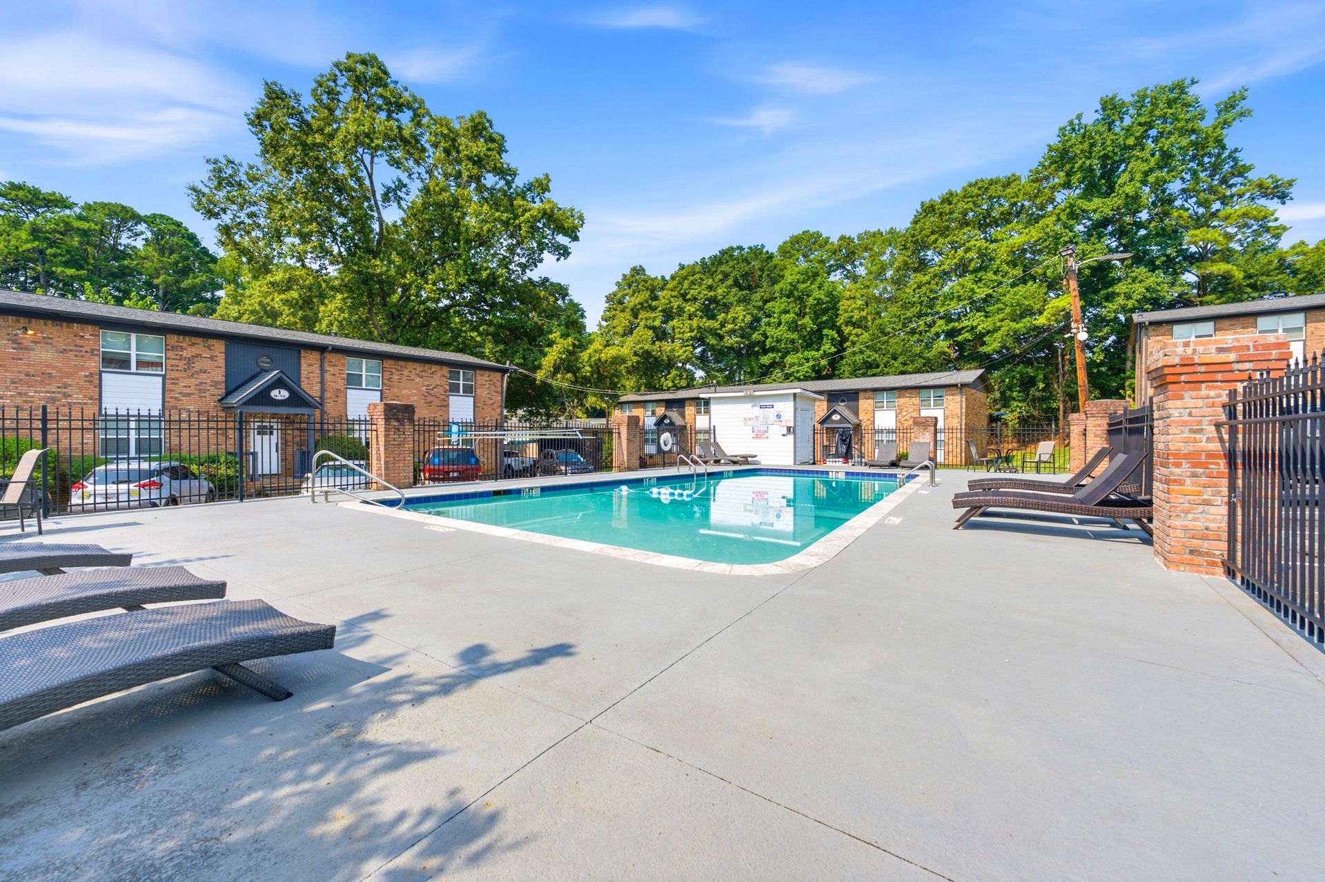 Outdoor apartment pool courtyard with lounge chairs, brick buildings, and trees under a clear blue sky