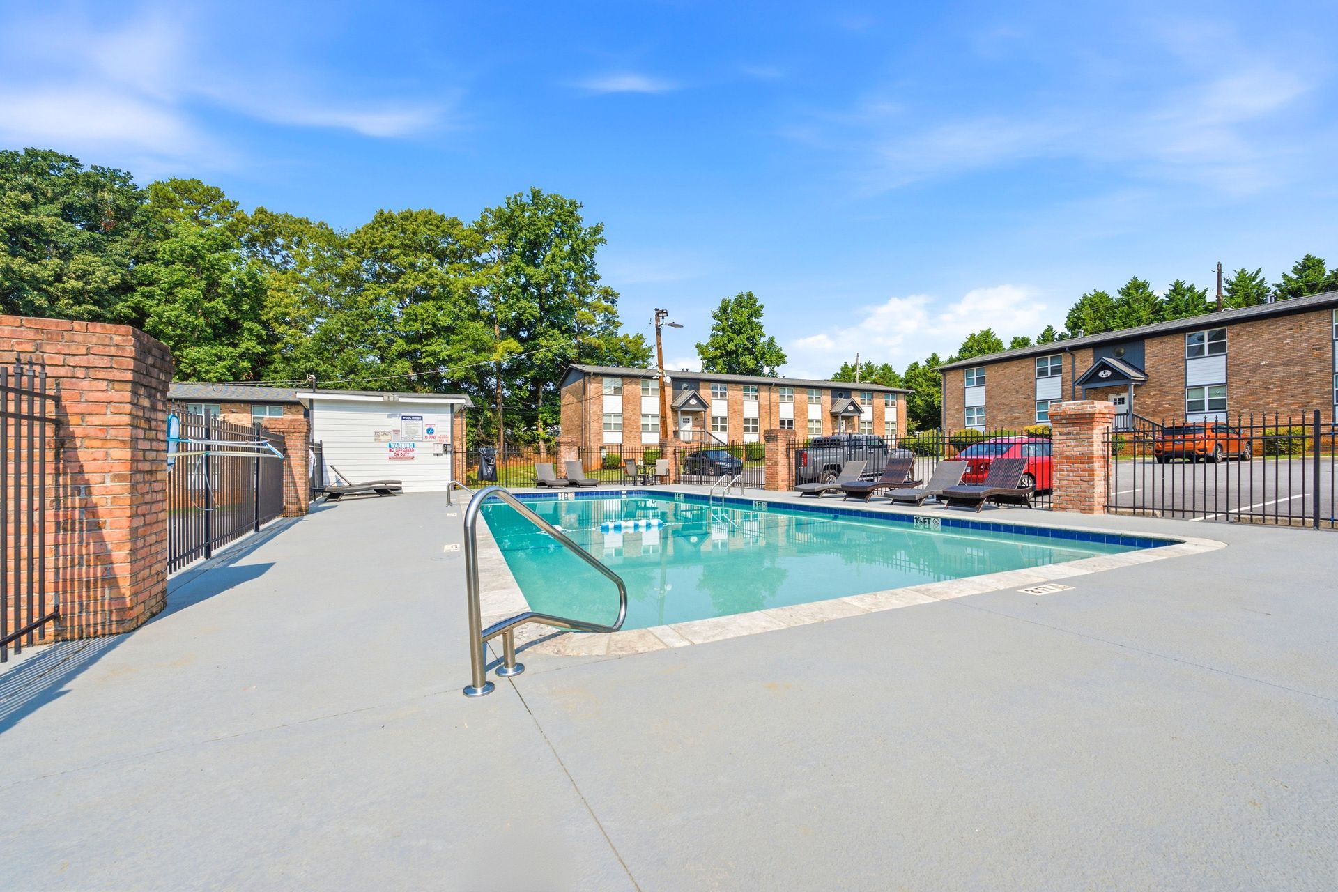 Outdoor apartment pool with lounge chairs, brick buildings, and trees on a sunny day