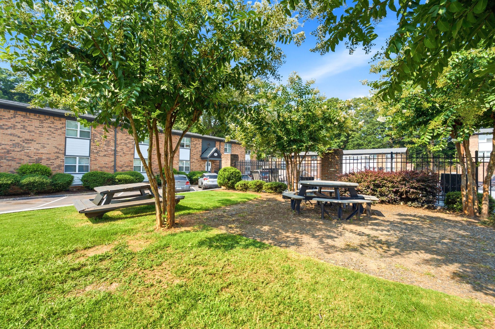 Shaded courtyard with picnic tables, trees, grass, and brick buildings under a blue sky