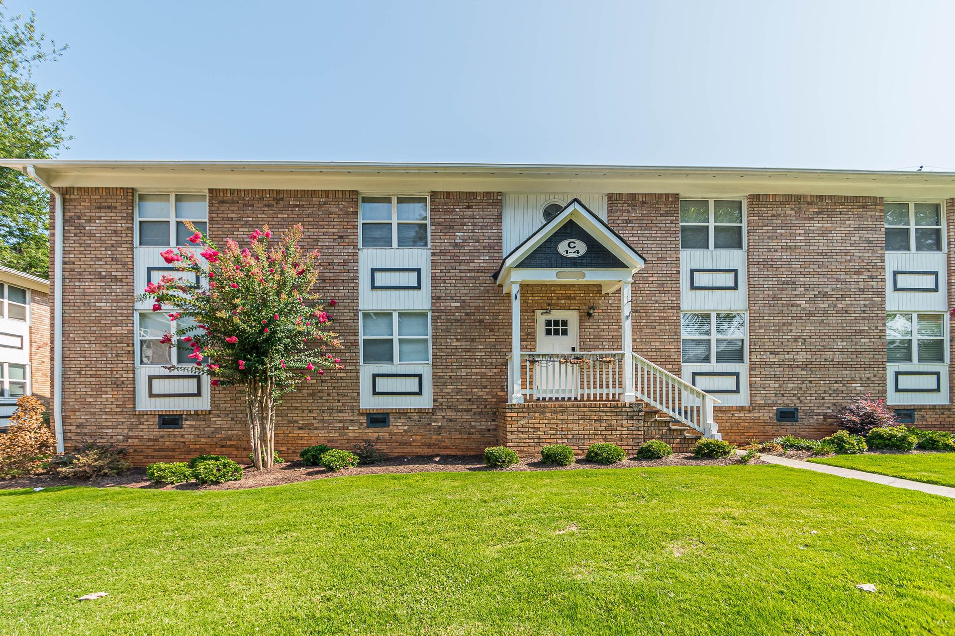 Single-story brick apartment building with a green lawn and small flowering tree in front