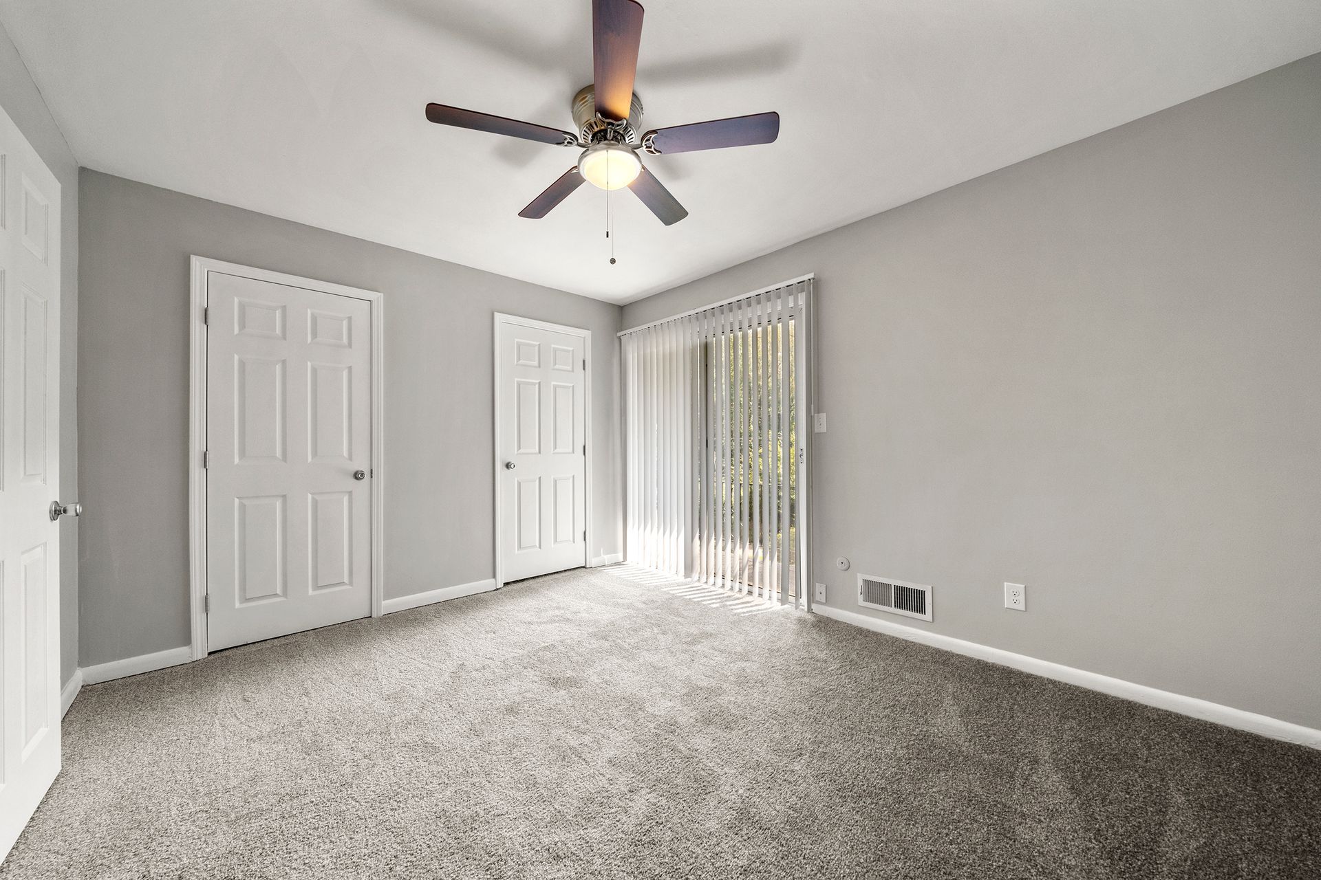 Empty gray bedroom with carpet, ceiling fan, two closet doors, and a sliding glass door with vertical blinds