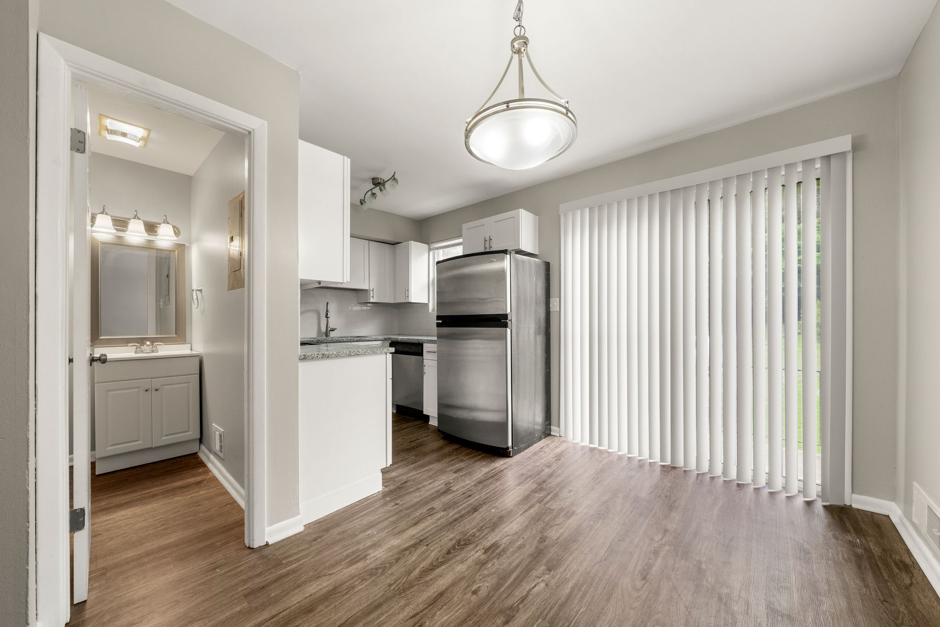 Bright modern kitchen with stainless steel fridge, white cabinets, wood floors, and vertical blinds