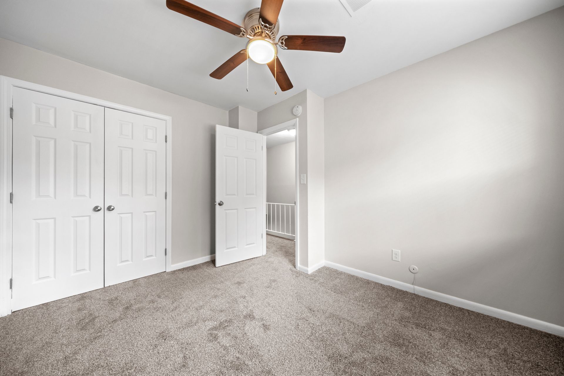 Empty beige bedroom with carpet, white closet doors, ceiling fan, and open door to hallway