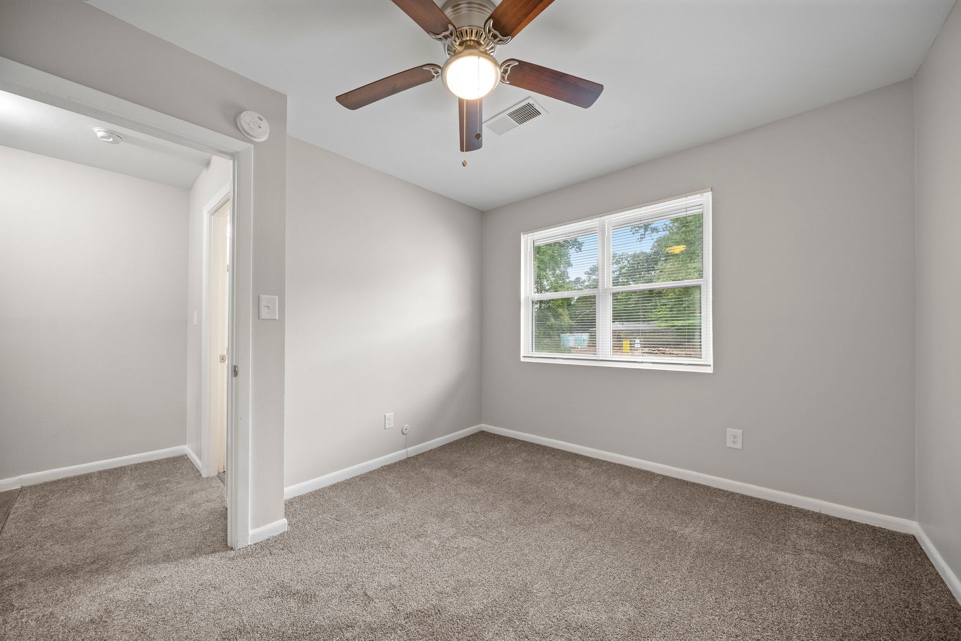 Empty carpeted bedroom with gray walls, ceiling fan, and a window letting in daylight