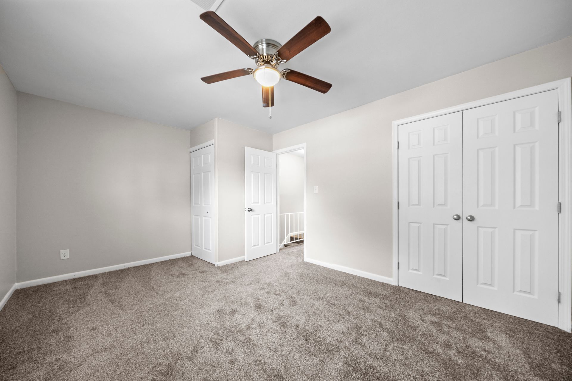 Empty bedroom with gray carpet, white walls, double closet doors, and a ceiling fan.